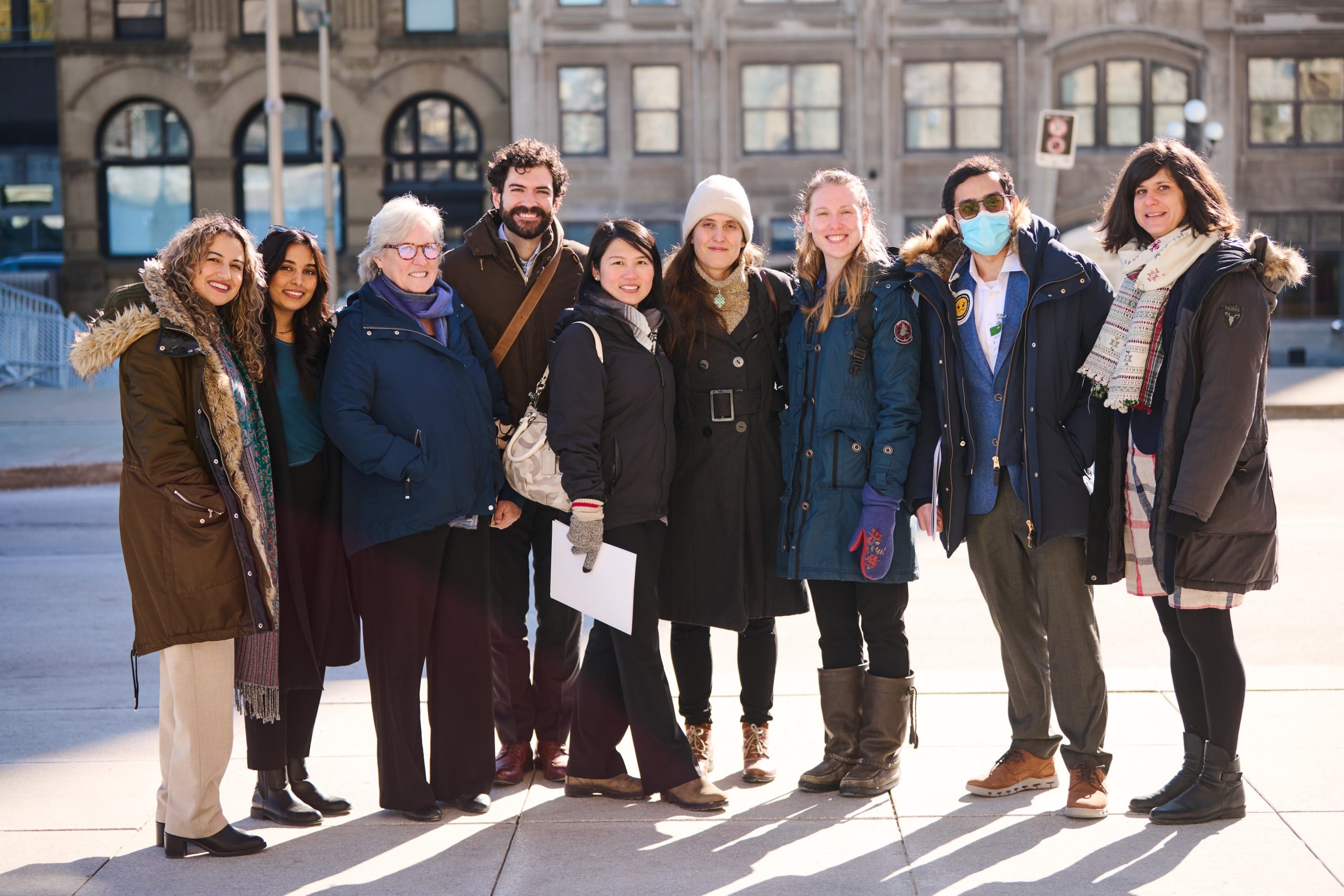 Seven health professionals and two CAPE stand together on a sunny February day in Ottawa with Parliament buildings in the background.